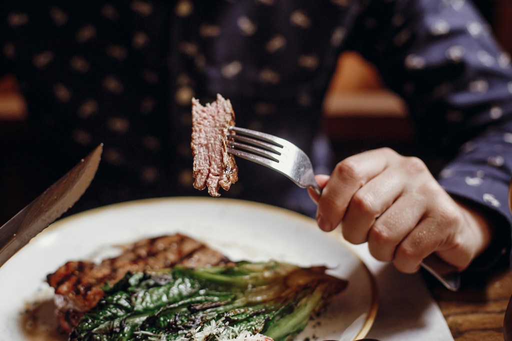 A person using a fork and a knife to eat a steak with grilled vegetables.