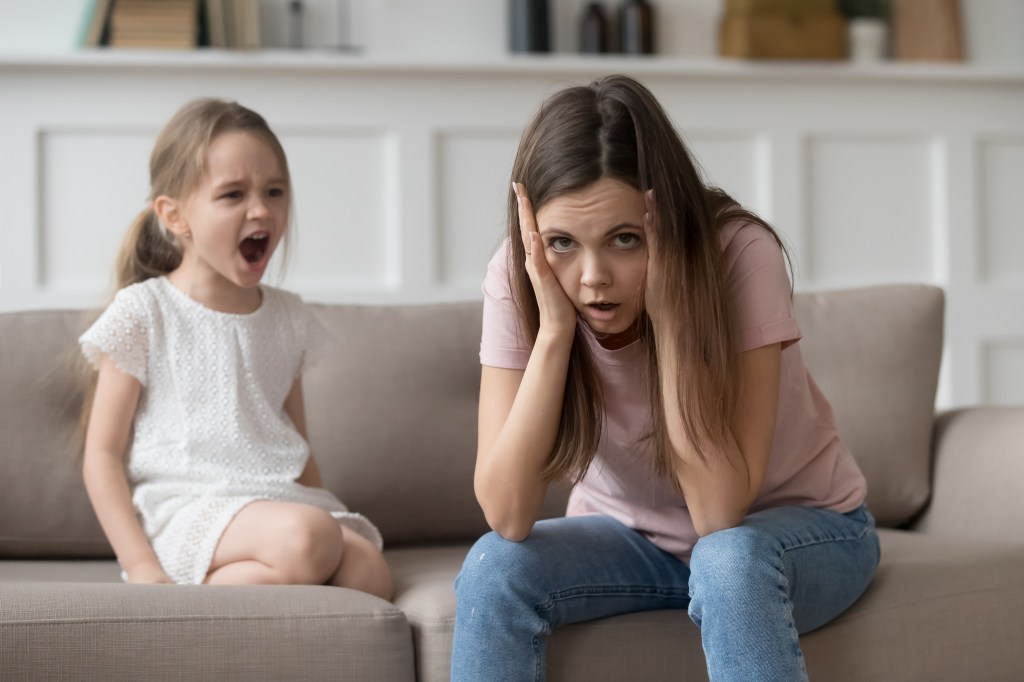 A stressed mother sits on a sofa next to her daughter shouting.