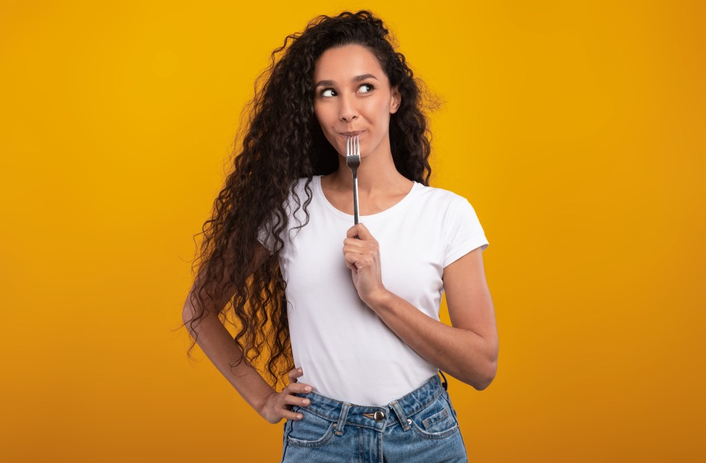 Woman who holds a fork in her mouth, looking thinking to one side.